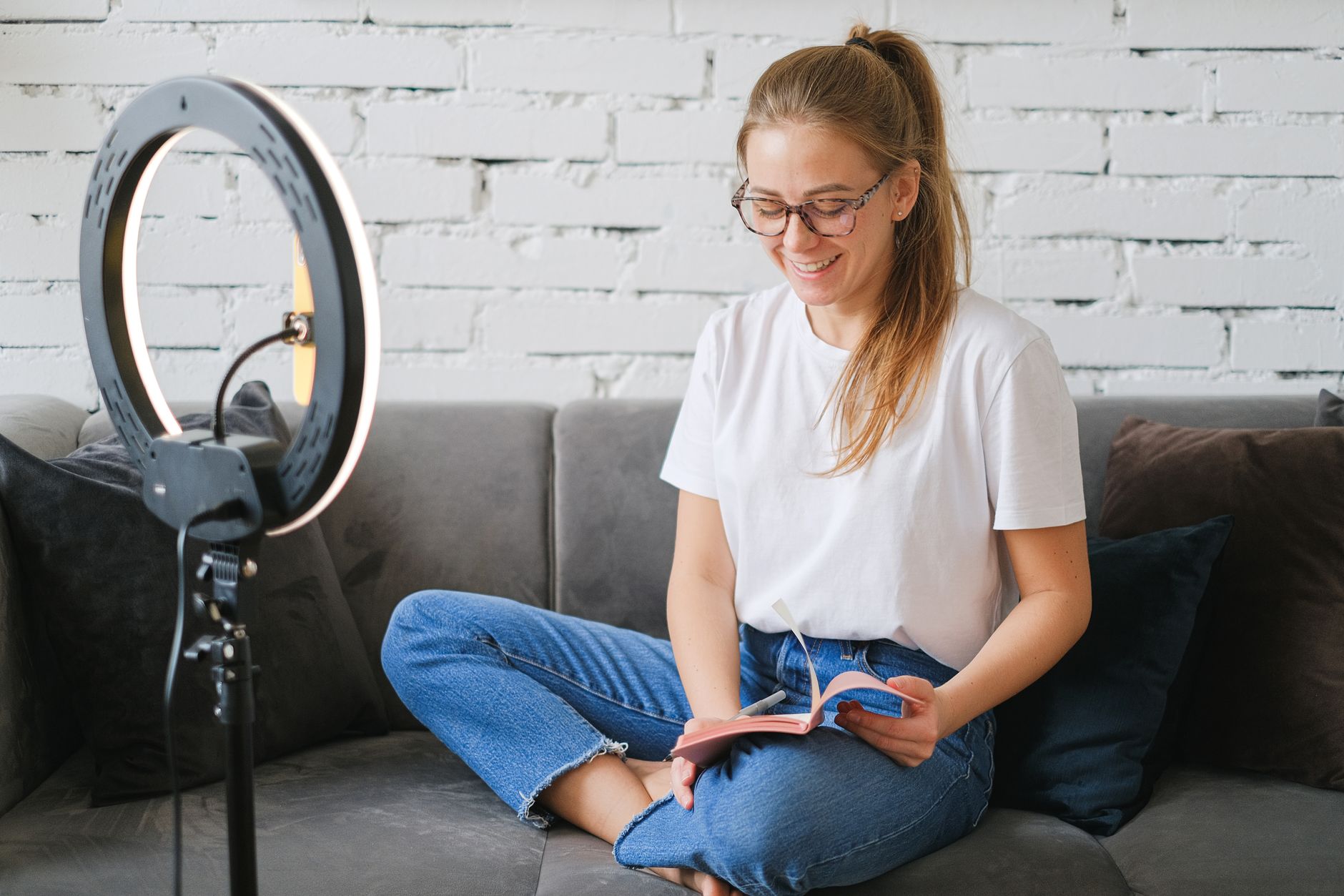 Smiling woman in a white T-shirt and jeans sitting cross-legged on a grey sofa at home, reading a notebook beside a ring light on a stand
