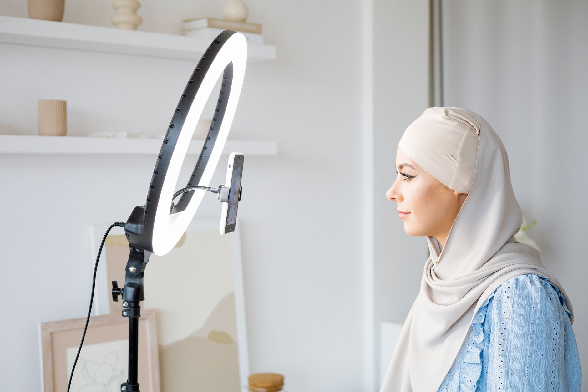 Woman in a cream hijab and blue blouse seated beside a ring light with a smartphone clipped to it, in a bright room at home