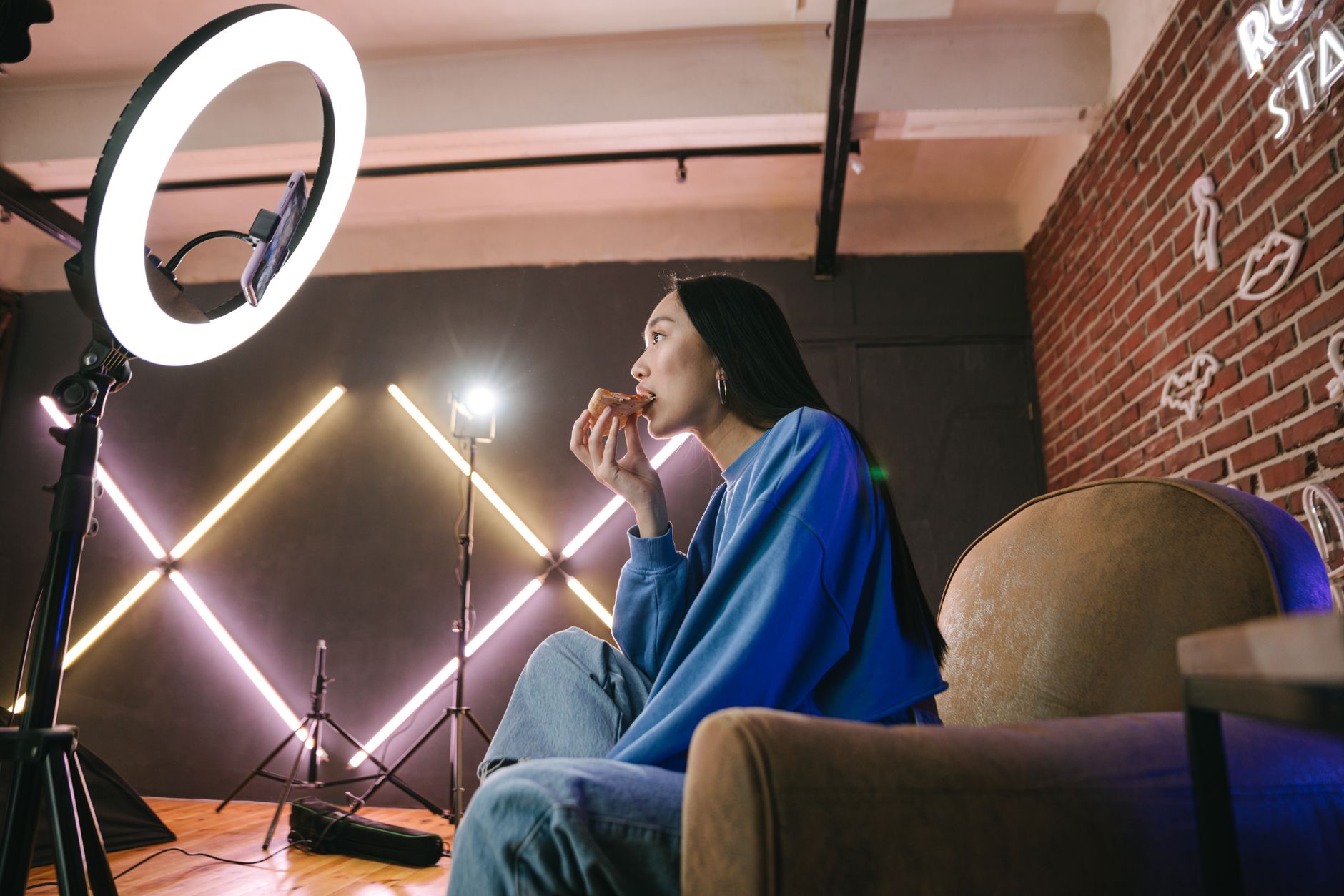 Woman in a blue sweatshirt eating a slice of pizza on camera while a phone on a ring-light stand records her